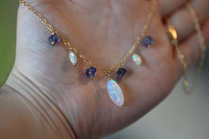 Gold necklace with opal and purple gemstones held in a hand against a blurred background. Layering necklace white Ethiopian opal with purple fluorite gemstones adjustable length jewelry OpalOra.