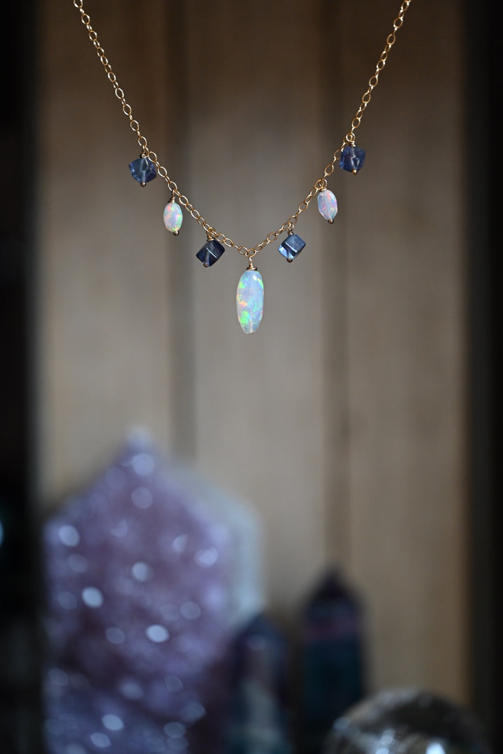 Delicate gold necklace with gemstones on a wooden surface. Natural white opal necklace featuring pink and blue flash with purple gemstone accents OpalOra Black Hills