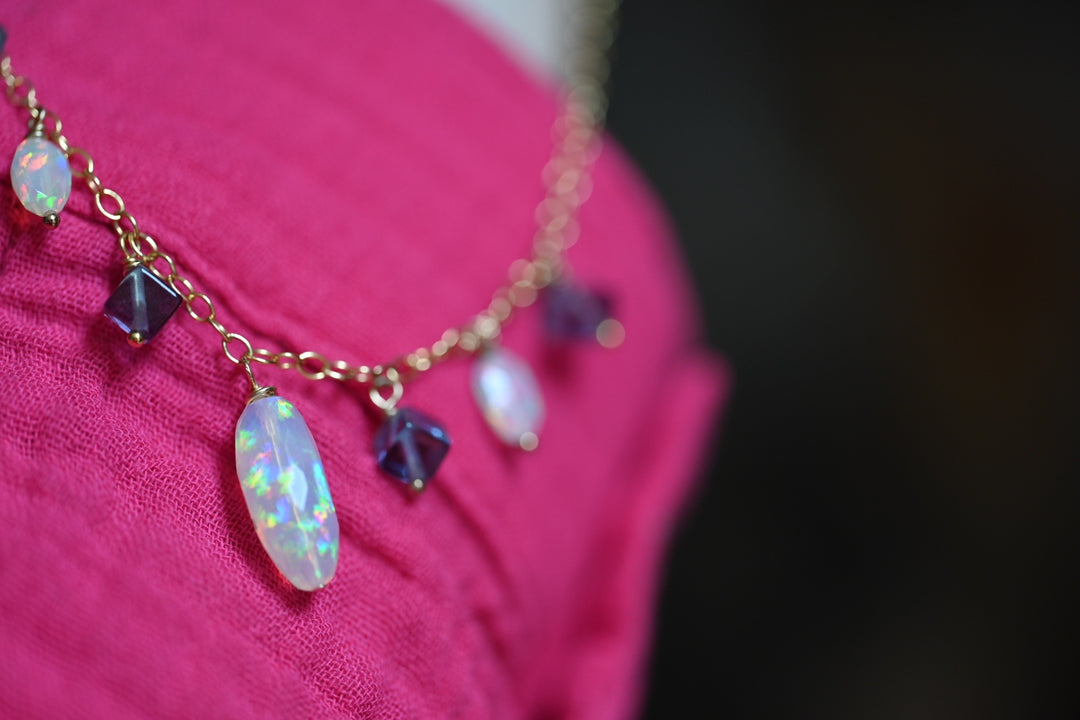 Necklace with opal and purple gemstones on a pink garment against a dark background. Women's opal necklace with color play flash purple fluorite beads 14K gold jewelry OpalOra Black Hills.
